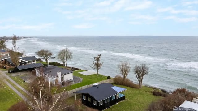 Black house stands still by the coast, surrounded by lawn and trees, with the sea rolling in the background under a slightly cloudy sky.