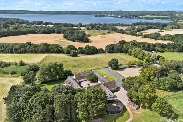 A white farmhouse with a thatched roof set amidst lush nature surrounded by forests and fields. In the background, a large lake and several green areas are visible.