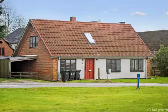 A brick house with a red roof located in a residential area. The house has a red door and a dormer. Three waste containers stand in front of the house.