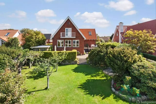 A red brick house with a balcony and garden in front. There are green trees and bushes in the garden. The sky is blue with scattered clouds.