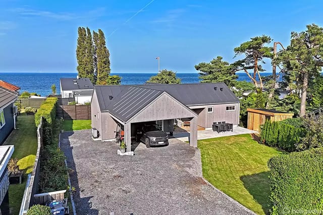 House with gray facade in front of a gravel area with a car parked under a carport. Background consists of lawn and sea view surrounded by trees and hedges.