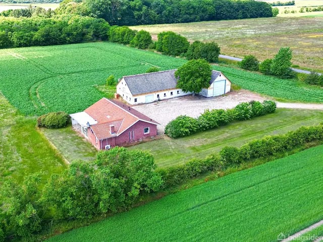 A red house with a flat roof next to a larger building in a green field. Trees surround the area, and a gravel road winds past.