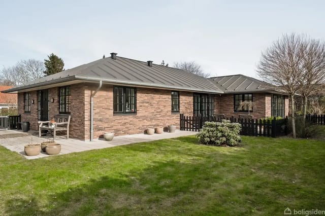 A brick villa with a black roof stands in a garden with a lawn and bushes. A fence surrounds the property, and some potted plants are on the terrace.