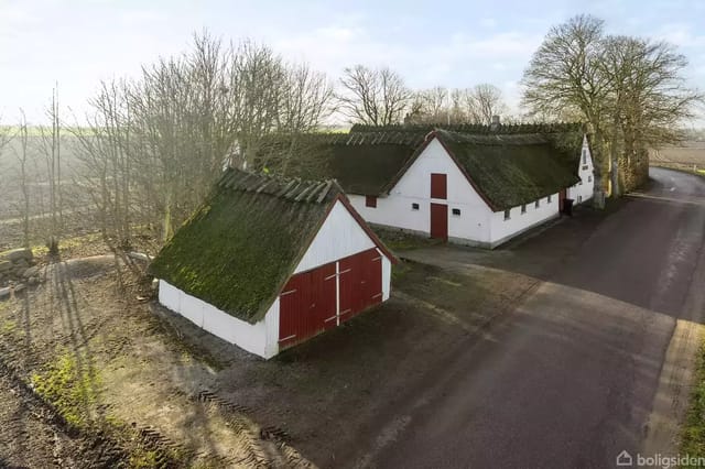 A traditional half-timbered house with a thatched roof located by a country road surrounded by trees and fields. A red door and a red shed are visible.