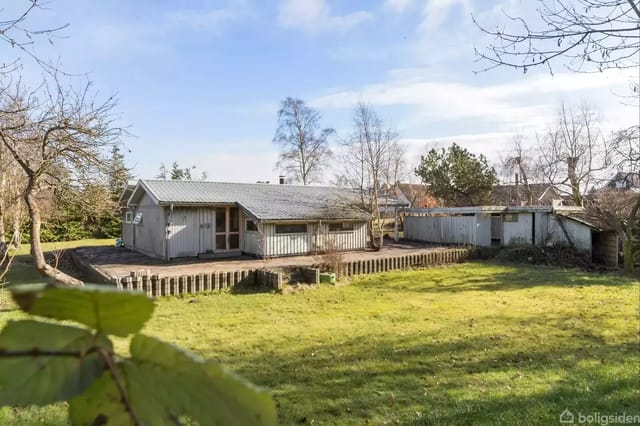 A small, gray wooden house stands quietly in a grass-covered garden surrounded by bare trees and bushes, with a clear sky in the background.