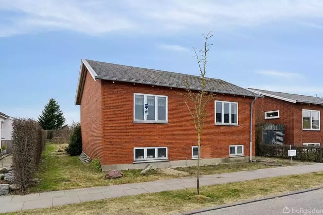 Red brick house on a lawn by the sidewalk. The stairs lead to an entrance. The background shows a residential area with blue sky.