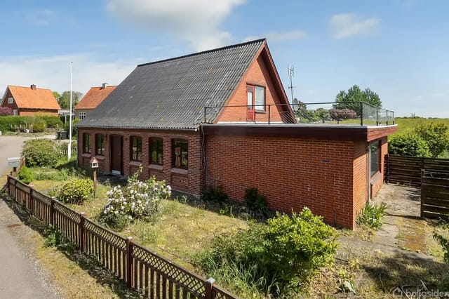 A red brick house with a sloped black roof stands in a small garden surrounded by green plants. The house has a rooftop terrace, and there are several houses in the background.