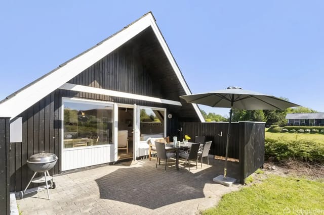 Black-painted holiday home with pitched roof, sunlit terrace, and outdoor dining area. Lawn and bushes surround the area.