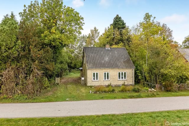 A small yellow house stands quietly among lush green trees. Gravel road in front of the house. The house has a black roof and three windows facing the road.