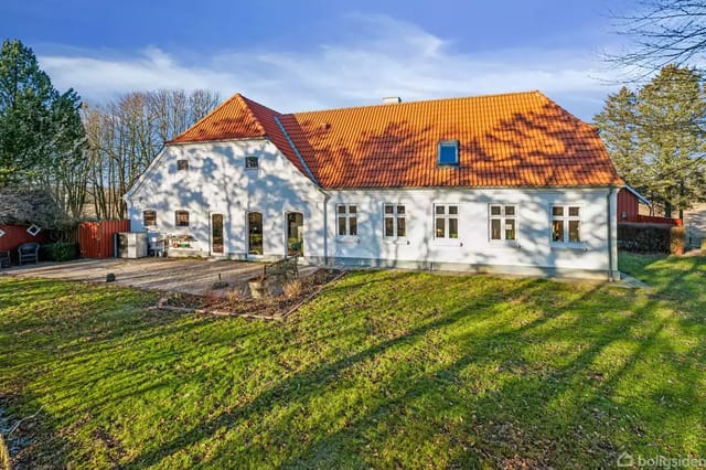 White building with red tile roof on a lawn, surrounded by trees and a clear blue sky. Concrete terrace in front of the building.