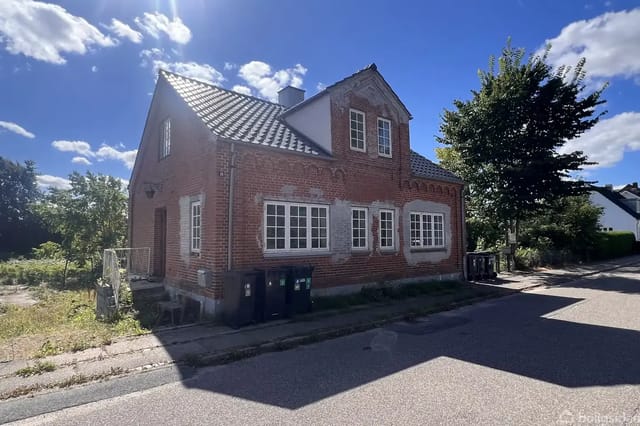 A red brick house stands quietly along a road on a sunny day, surrounded by trees and a lawn. There are two stories and large windows.
