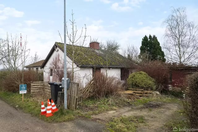 A small, white house with a moss-covered roof stands in an overgrown garden. Traffic cones and a sign with the text 'For sale' are seen by the road in front of the house.