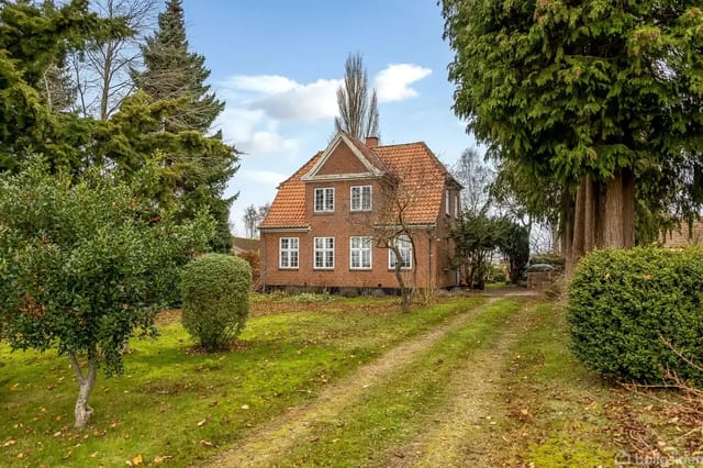 Red brick house with white windows, set on a gravel road surrounded by lush green vegetation and large trees under a blue sky with clouds.