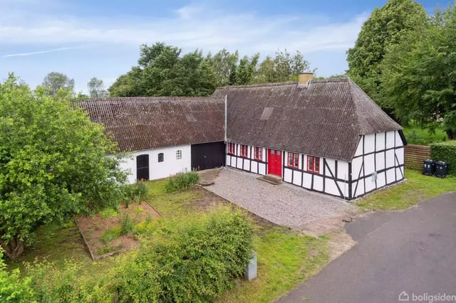 Half-timbered house with thatched roof and red door in a lush garden with trees around. A paved road runs in front of the house.