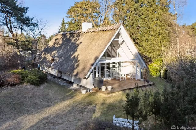 Thatched house standing quietly surrounded by a green garden and tall trees. Large glass sections and terrace in front of the house. Clear blue sky above.