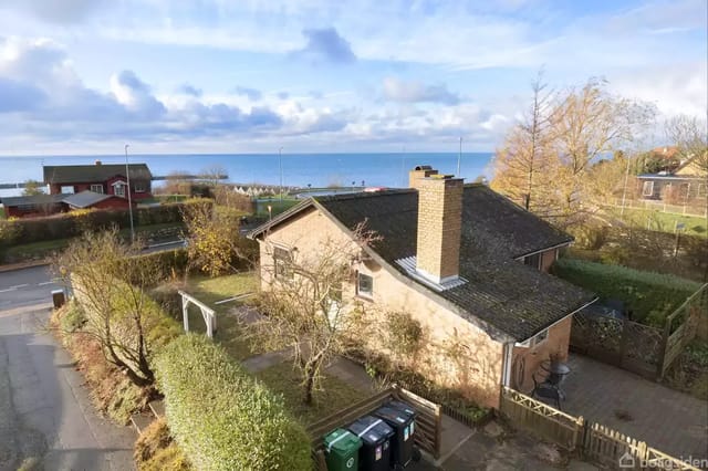 House with gray roof seen from above surrounded by trees and bushes. In the background, there is a view of the sea under a partly cloudy sky.