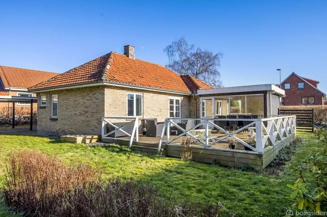 A brick house with a red tiled roof has a large wooden terrace surrounded by a white railing. Lawn and bushes adorn the garden, while a clear blue sky forms the background.