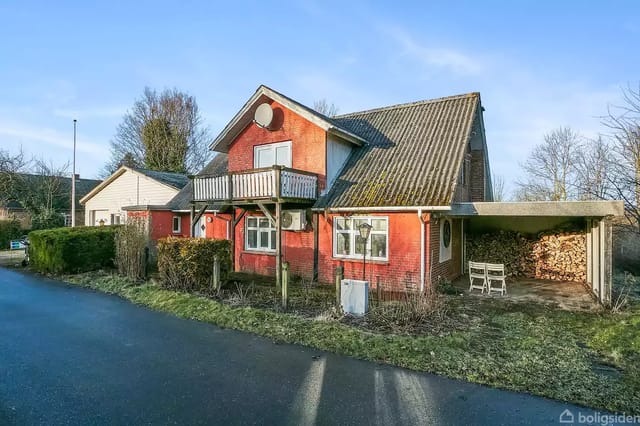 A red brick house with a balcony and sloped roof stands next to a road. Next to the house is a carport with wood stacks and a chair.