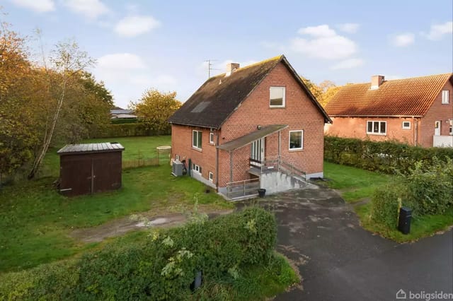 A red brick house surrounded by a green garden with a shed to the left. The house has a sloping roof, and the entrance is connected with a small staircase. The weather is clear.