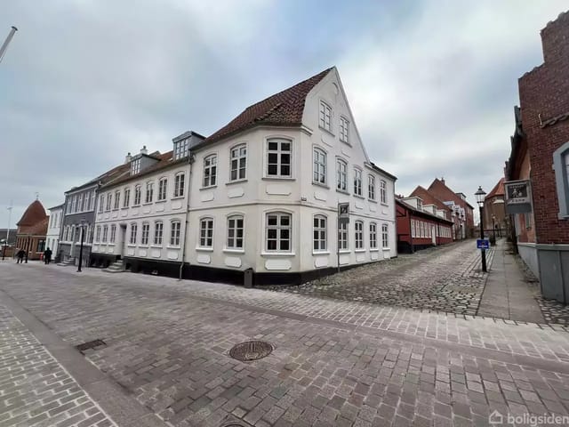 White, two-story house with red tile roof on a cobblestone street corner in a quiet urban environment. English sign on the facade: 'Old Nytorv Antiques'.