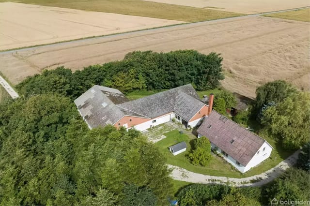 Building with sloping roof surrounded by trees, seen from the air. Surrounding fields stretch into the distance, creating a peaceful, rural atmosphere.