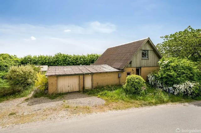 A classic older house with a sloped roof, surrounded by green bushes and trees, with a gravel driveway in front.