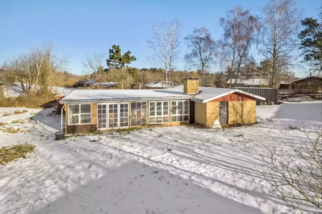 A simple house with large windows stands on a snow-covered plot. The house has a chimney and is surrounded by trees and scattered bushes in a rural area.