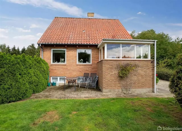 Red brick house with red tile roof in a garden with green lawn and paved terrace. A glazed extension and garden furniture are seen next to a green bush.