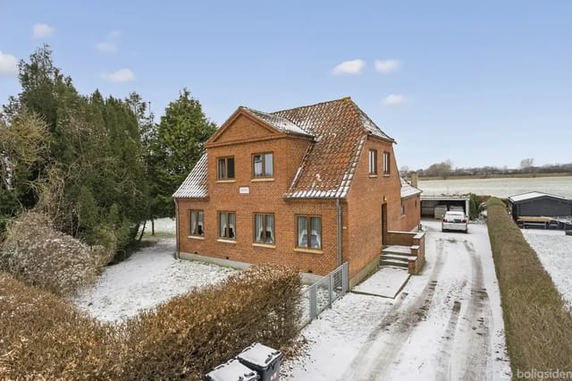 A red brick house stands quietly in a snow-covered landscape, surrounded by hedges and trees. A snow-covered driveway leads to a garage in the background.
