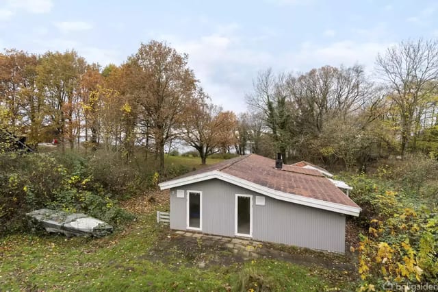 A gray, wood-clad holiday home surrounded by trees in autumn colors. A boat lies on the grass to the left. The sky is overcast.