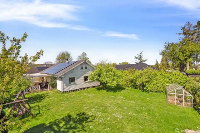 A white house with solar panels on the roof stands in a sunny garden with a swing to the left and a small greenhouse to the right, surrounded by green trees.
