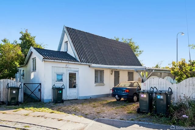 The house with a car parked in front, surrounded by waste containers and fencing. The facade is white with a black roof, and there are a few bushes and trees in the background.