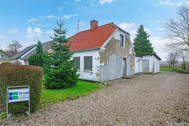 A house with a red tile roof stands by a gravel road. In front are two large pine trees, and to the left is a grassy area with a 'For Sale' sign from Nybolig.