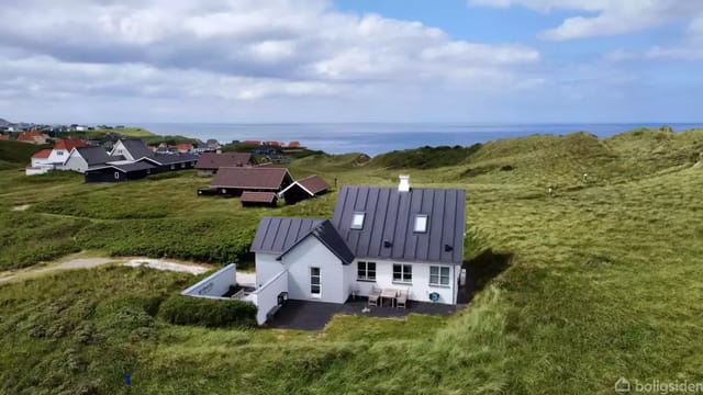 A white house with a black roof stands quietly surrounded by green hills and grass. In the background, more houses and the sea can be seen under a blue sky with clouds.